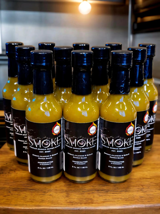 Twelve 5 oz glass bottles of Smoke Handcrafted Smoked Jalapeno and Garlic Pepper Sauce arranged in rows on a wooden counter under warm, bright overhead lights.