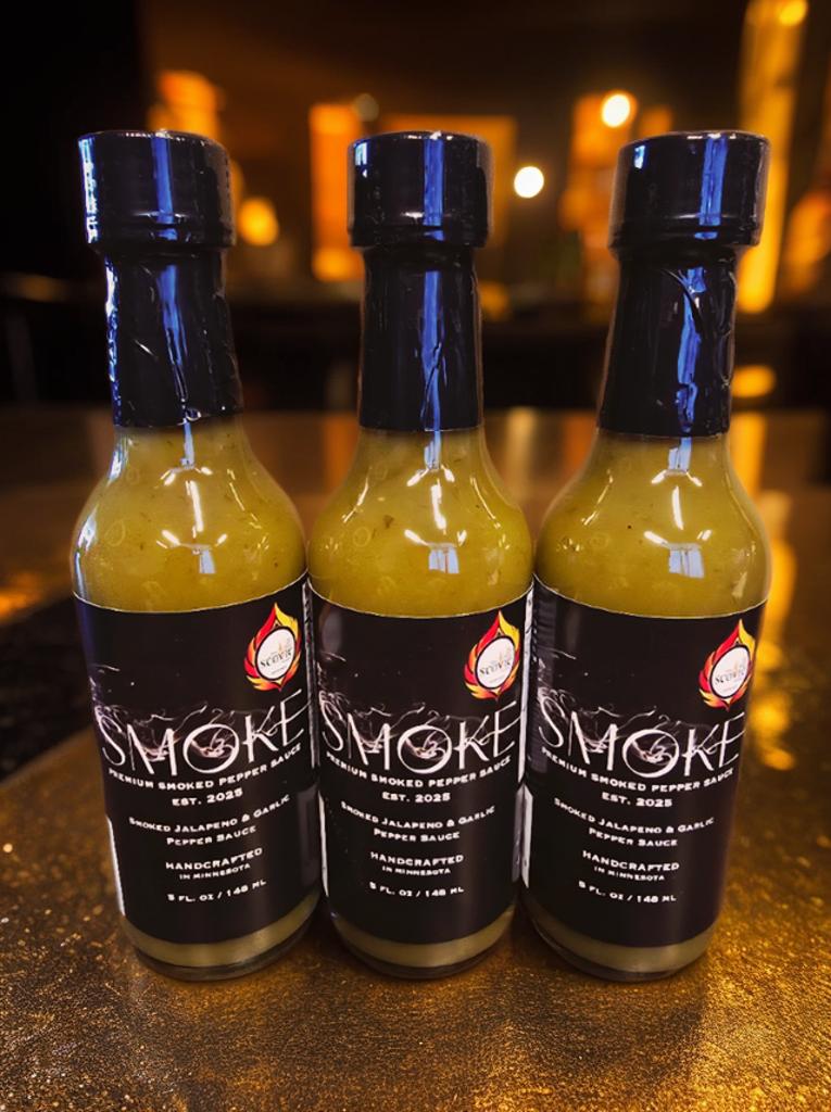 Three 5 oz glass bottles of Smoke Premium Smoked Pepper Sauce lined up on a counter, featuring a sleek black label with a white logo and text detailing a handcrafted smoked jalapeno and garlic flavor blend.