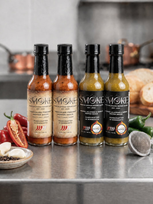 Four glass bottles of "SMOKE" brand premium smoked pepper sauce are arranged n a row on metallic kitchen countertop. The two bottles on the left feature tan labels with red and brown text reading "Freckled Fresno" Pepper Sauce.
The two bottles on the right feature black labels with white text reading "SMOKED JALAPENO & GARLIC PEPPER SAUCE"
Each label includes "Est. 2025" and "Handcrafted in Minnesota"
The background shows a softly blurred rustic kitchen environment with fresh peppers and ingredients.