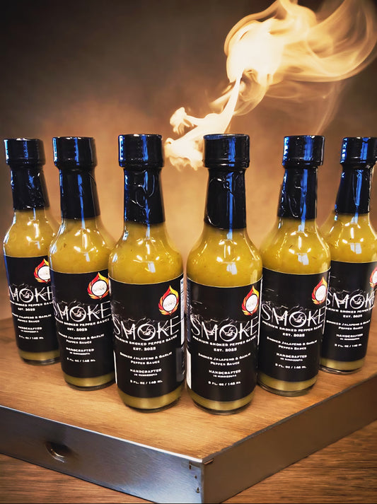 Six 5 oz glass bottles of Smoke Handcrafted Smoked Jalapeno & Garlic Pepper Sauce lined up on a rustic wooden stand with smoke rising in the background.